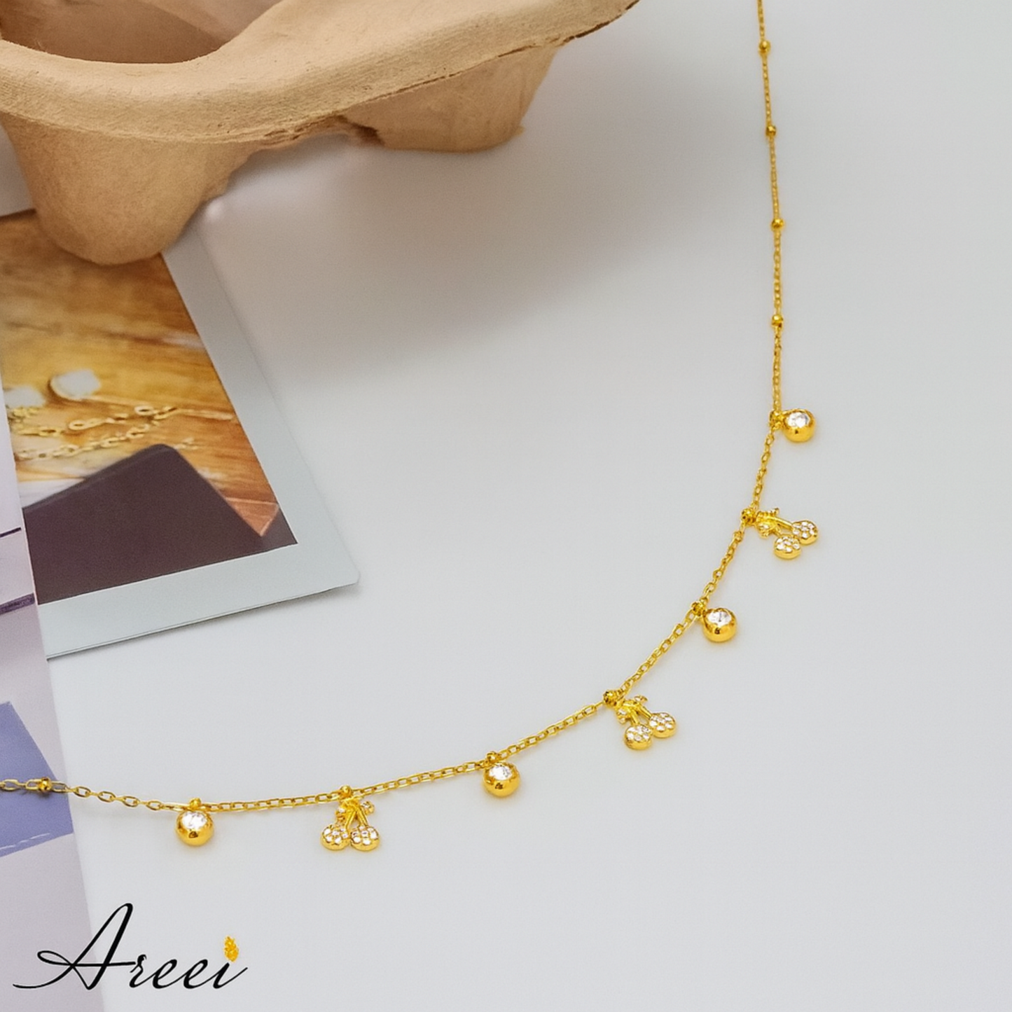 Gold necklace with small charms on a light surface, featuring the brand 'Areei'.