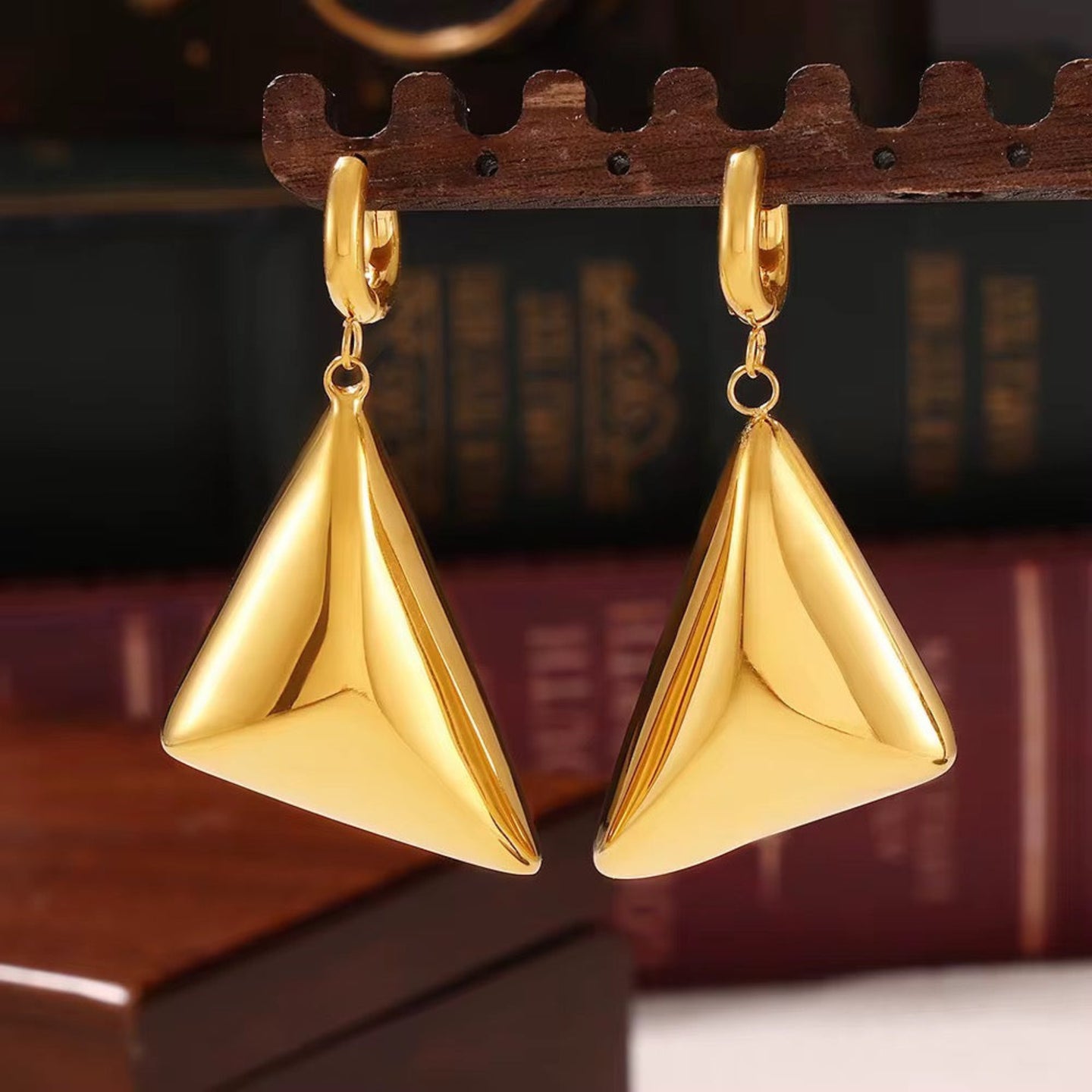 Gold triangle earrings on a wooden stand with a blurred bookshelf background
