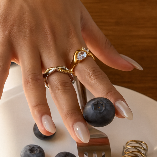 Hand with gold rings holding a blueberry on a white plate with a fork