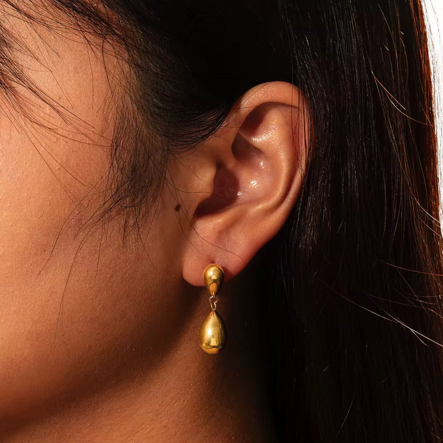 Close-up of an ear wearing a gold teardrop earring with dark hair and skin background.
