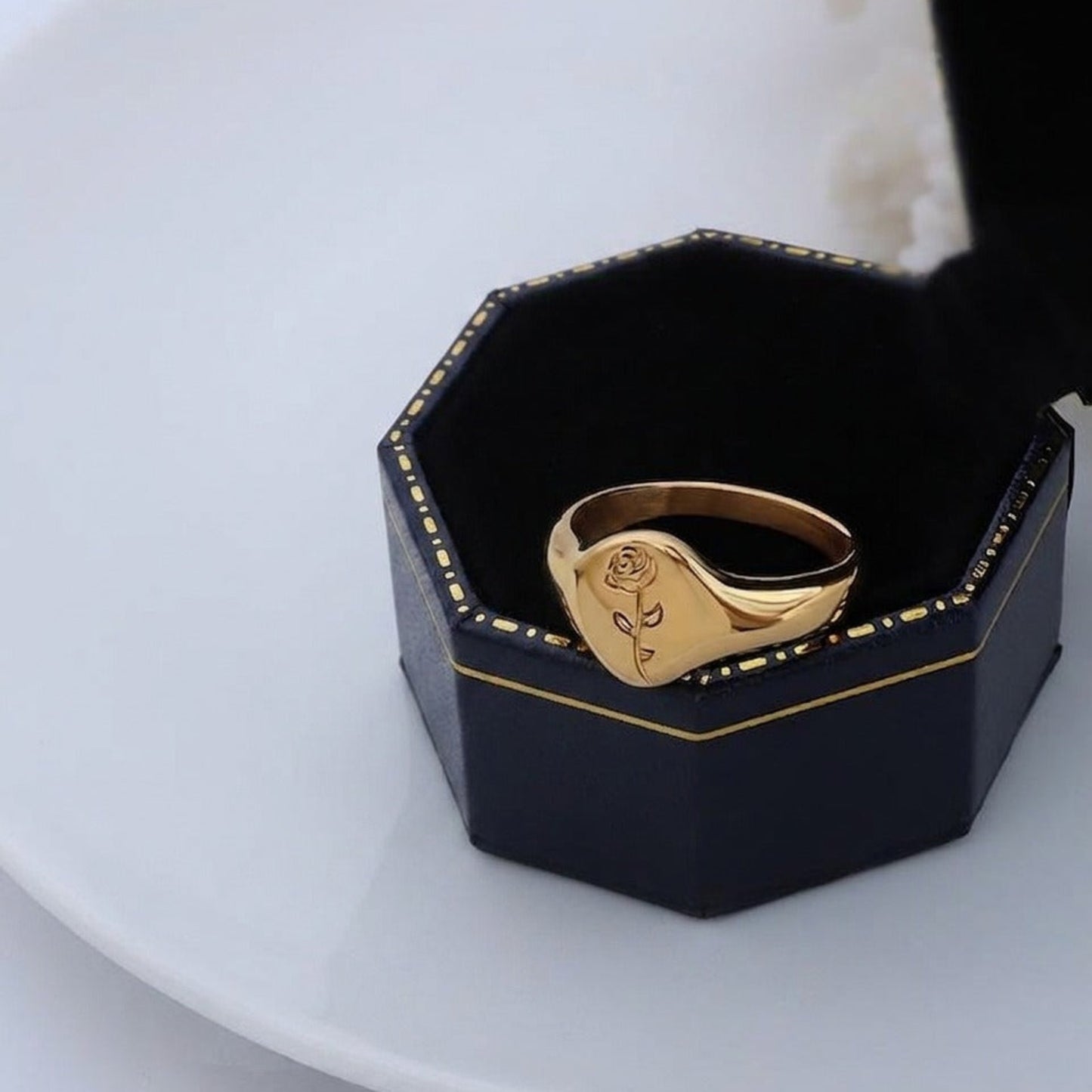Gold ring with rose design in a black hexagonal box on a white plate