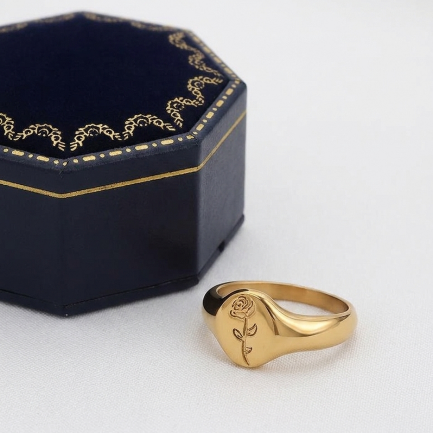 Gold ring with rose engraving on a white surface next to an ornate black box.