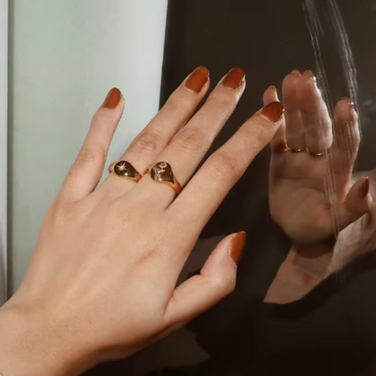 Hand with gold rings and brown nail polish against a neutral background