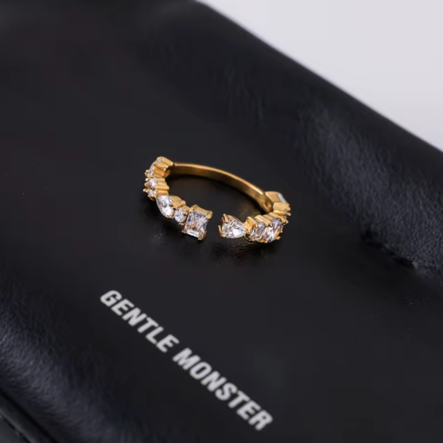 Gold hoop earring with clear stones on a black surface with 'Gentle Monster' branding.