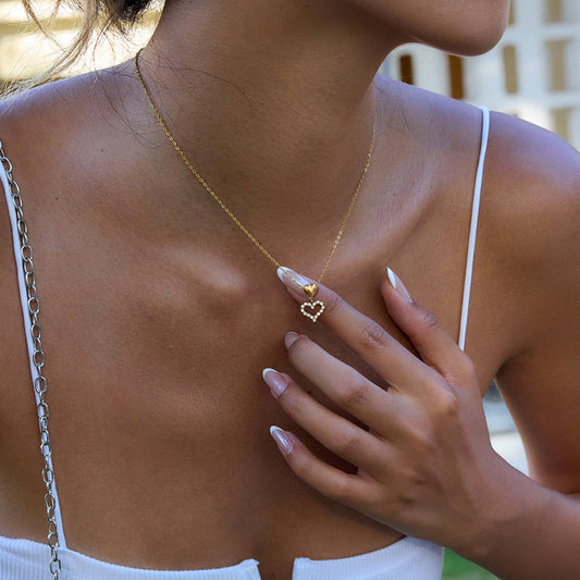 Woman wearing a gold necklace with a heart pendant, touching her chest.